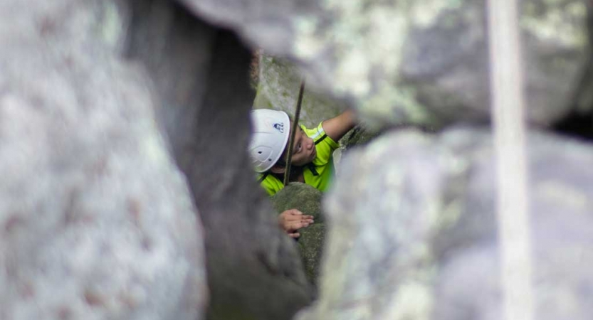 From above and through a crack in rocks, a person wearing safety gear concentrates as they rock climb.